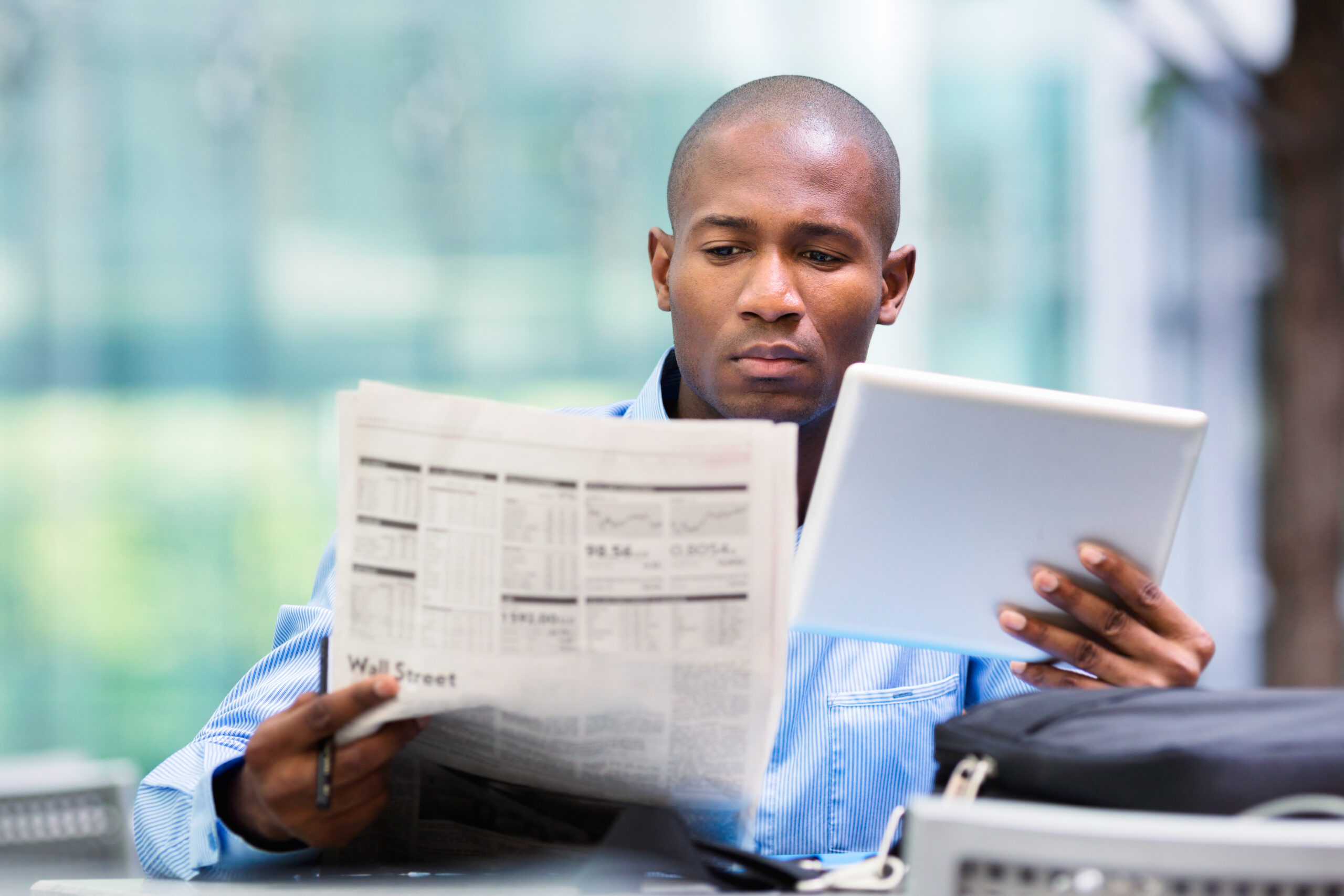 African American businessman comparing stock market quotes on his tablet and in the newspaper.