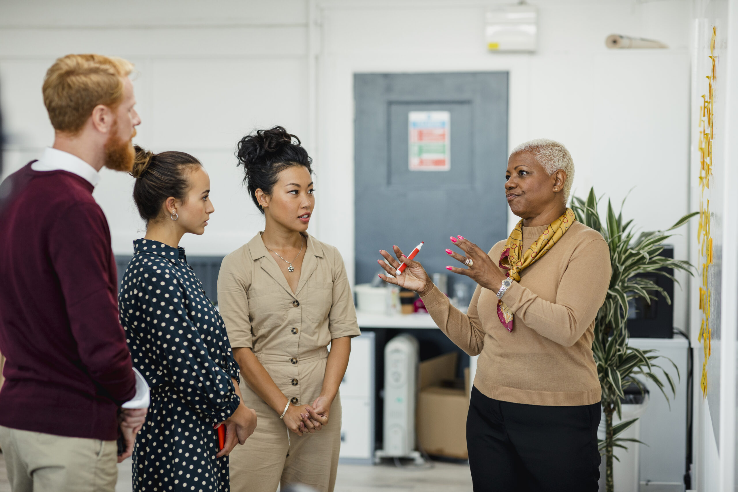 Colleagues standing in a small group discussing something while one women gestures with her hands holding a white board pen. They are standing in front of a whiteboard.